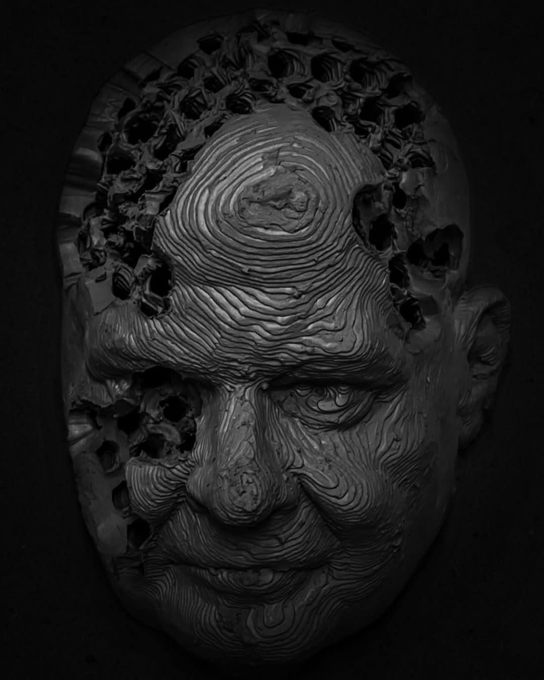 3D printed object by Anthony Bennett, a man's face in black clay with contoured surface and holes like a beehive.