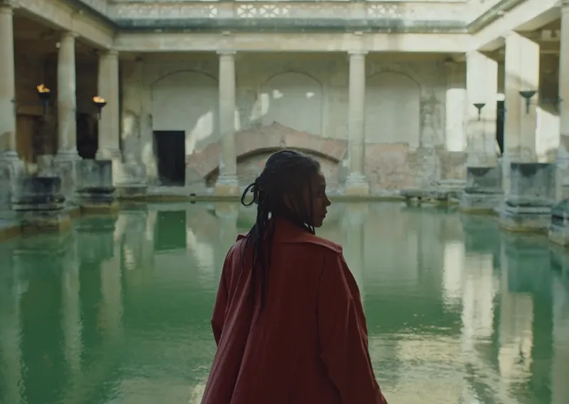 A woman with her back to the camera, looking at an ancient Roman bath with columns raising from a blue pool.