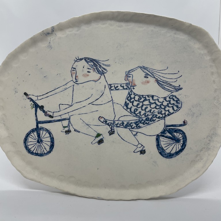 White porcelain plate by Becca Brown, featuring whimsical illustration of two people riding a tandem bike in blue ink.