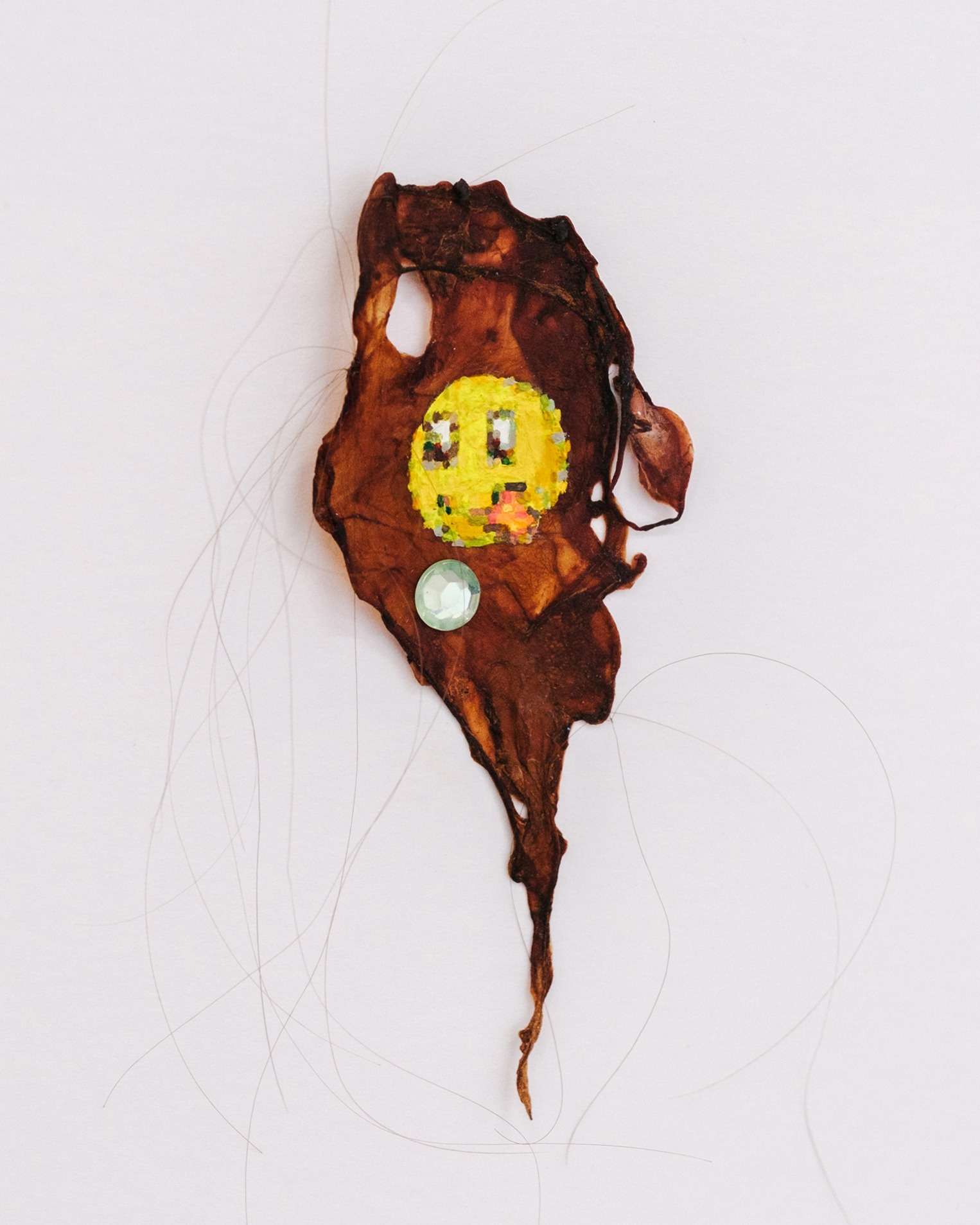 Artwork by Cat Brierley, a pixellated smiley face sticking out its tongue, painted on a scrap of brown organic material, with added gemstone and hair strands.