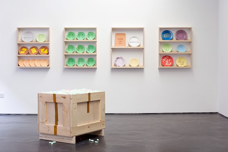 Installation by Charlotte Dawson featuring brightly coloured plates on wooden shelves, in the foreground is a wooden crate filled with green packing peanuts.