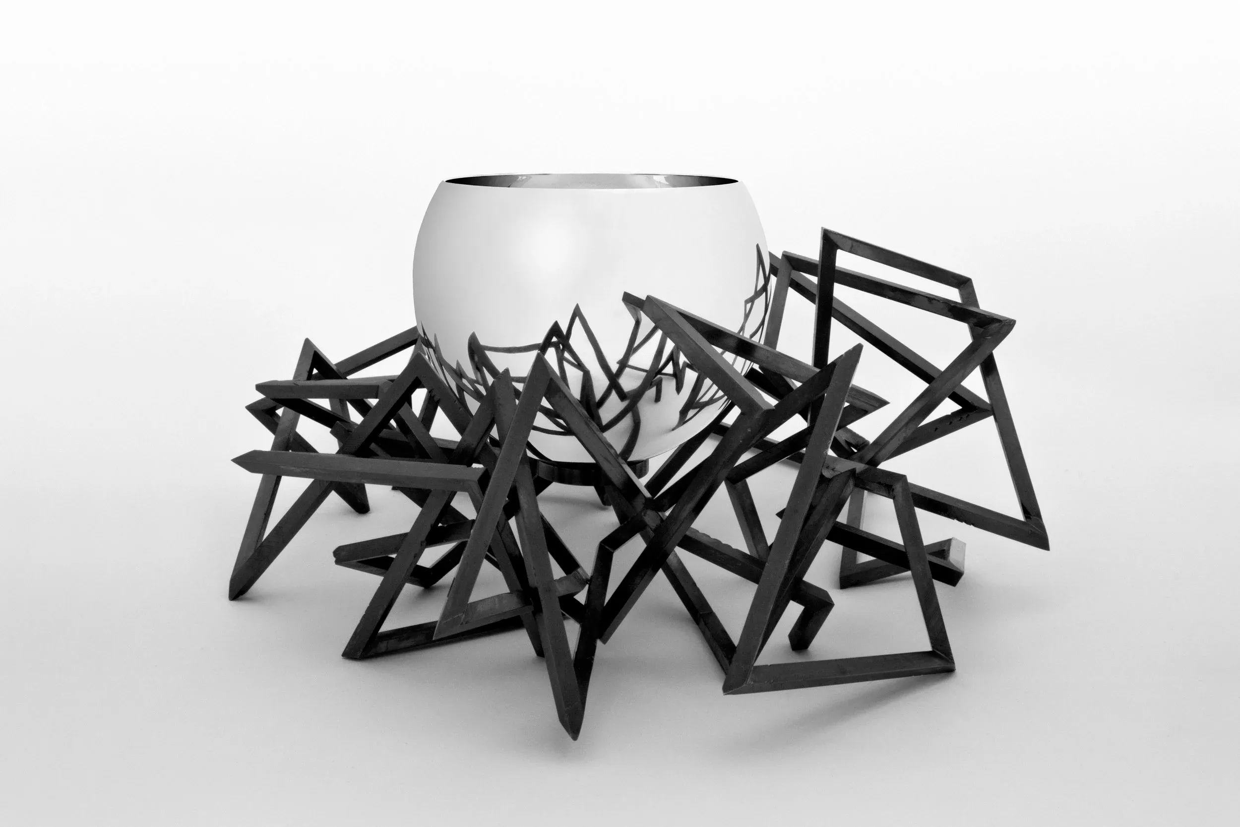 Silver bowl by Chris Knight, sat in a black geometric nest-like structure, which is reflected in the surface of the bowl.