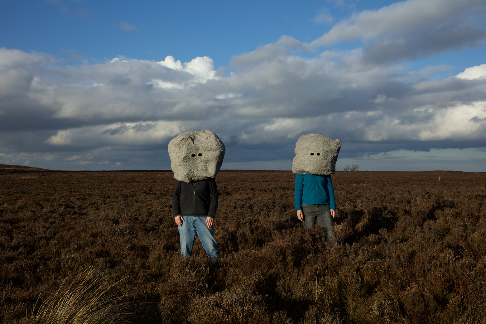 Photograph by Conroy / Sanderson, two people standing in a remote grassland. Their heads are covered by large boulders with eye holes.