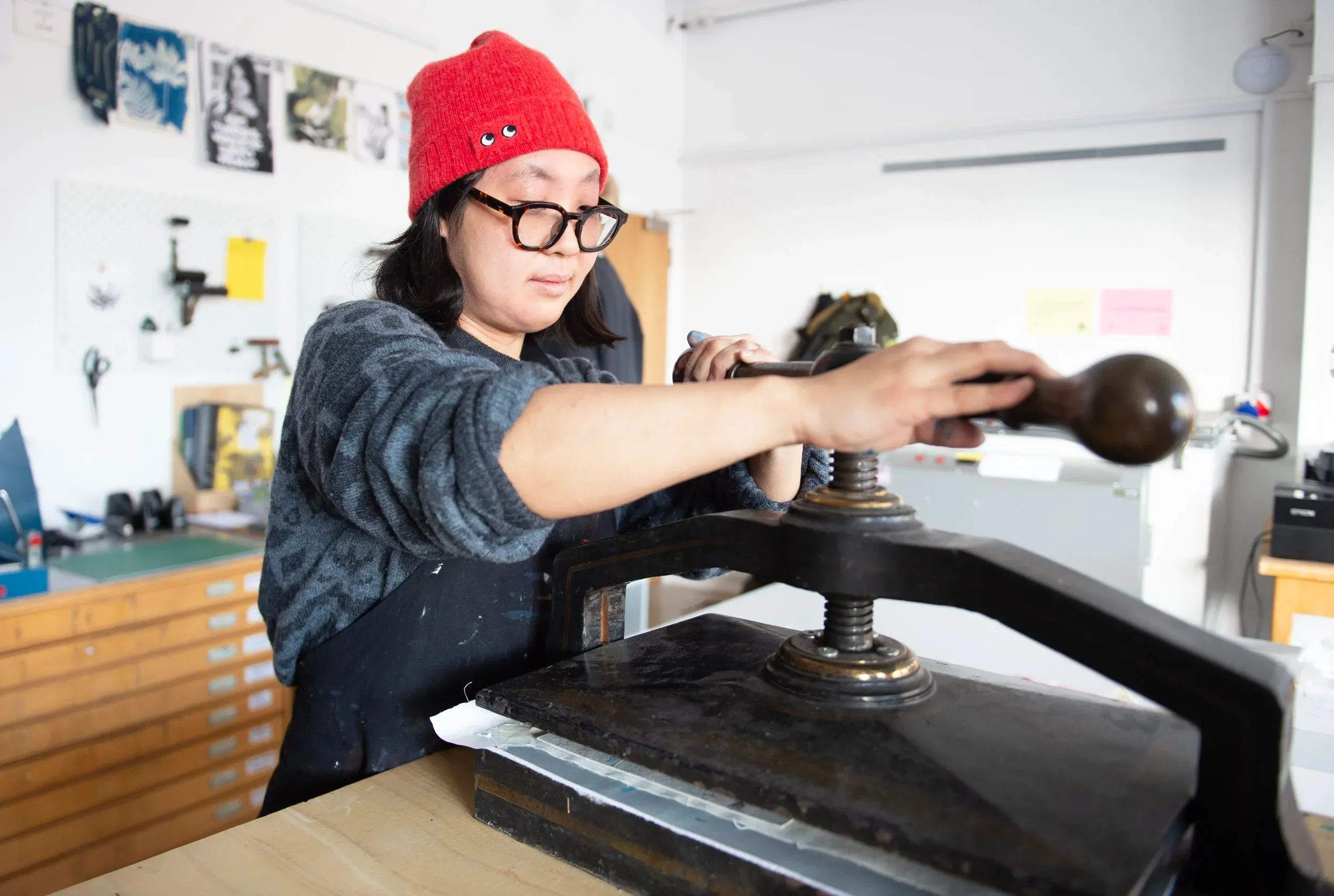 Woman wearing a red beanie, grey jumper, and glasses, twisting the handle on a printing press in Carousel Print Studio.