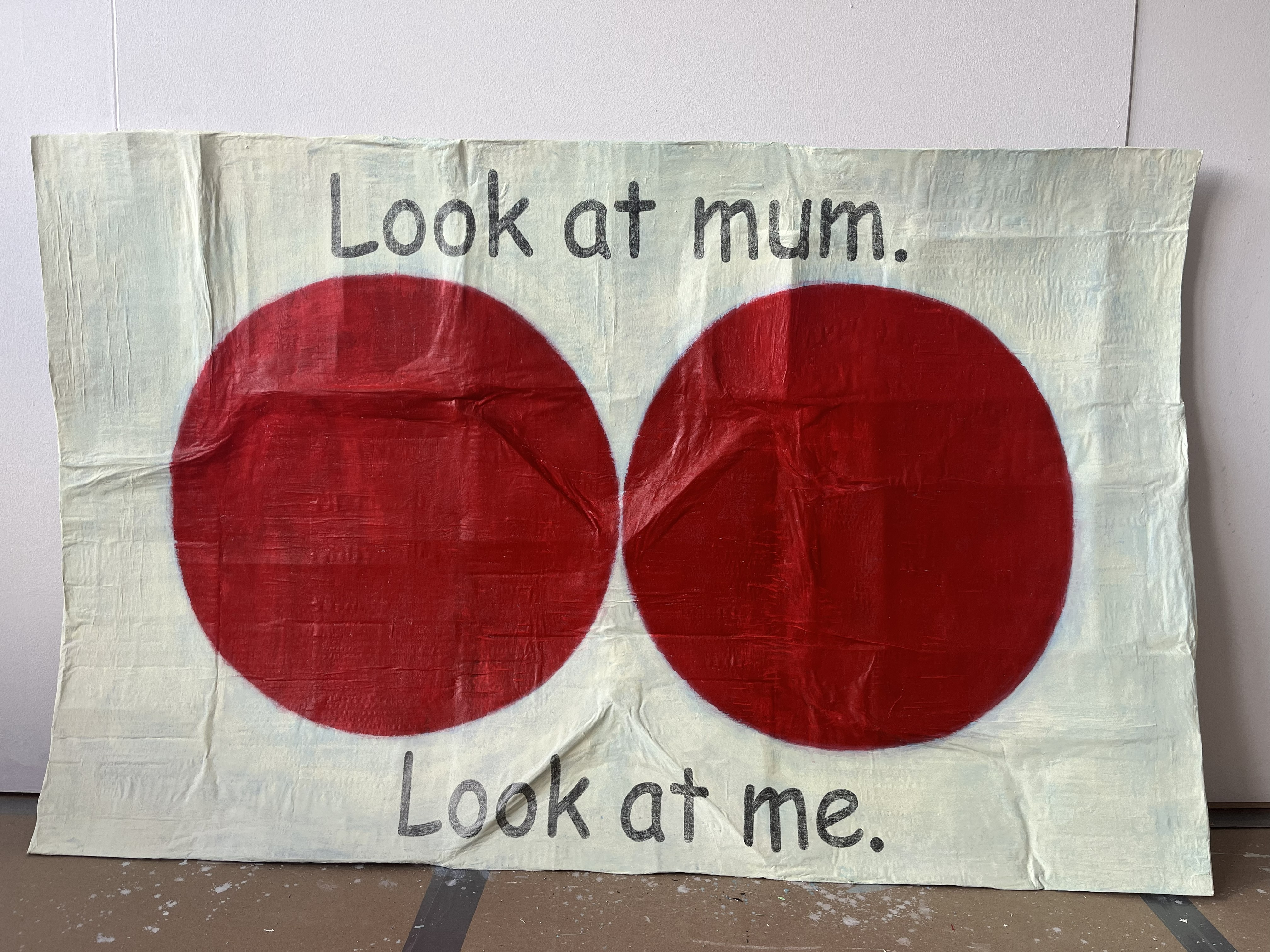 Crumped and creased piece of cardboard, painted pale yellow with two dark red circles in the centre, almost touching. Text reads "Look at mum. Look at me."
