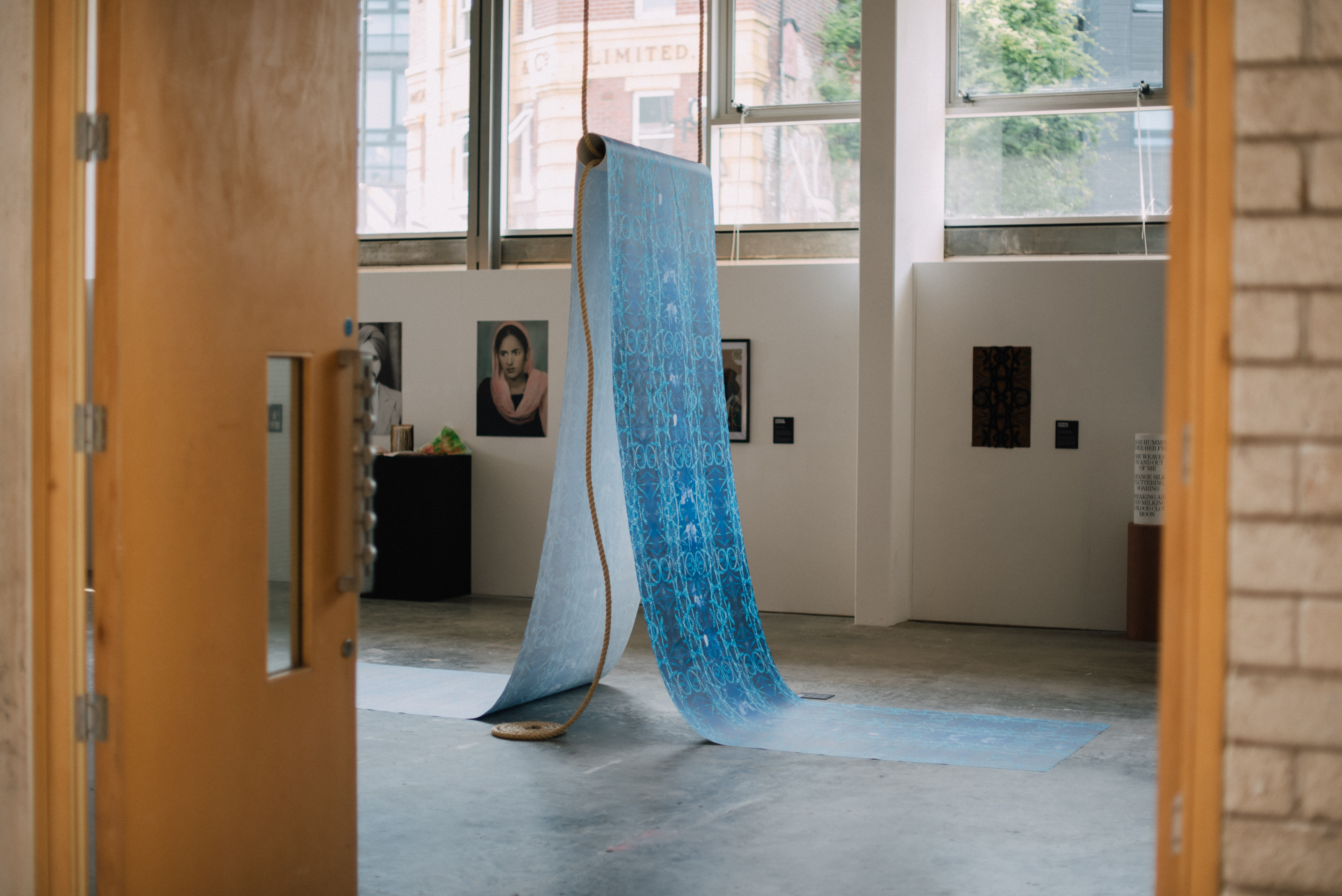 Shot peering through a doorway into a gallery space, exhibition featuring a large roll of blue patterened paper suspended from the ceiling by thick ropes.