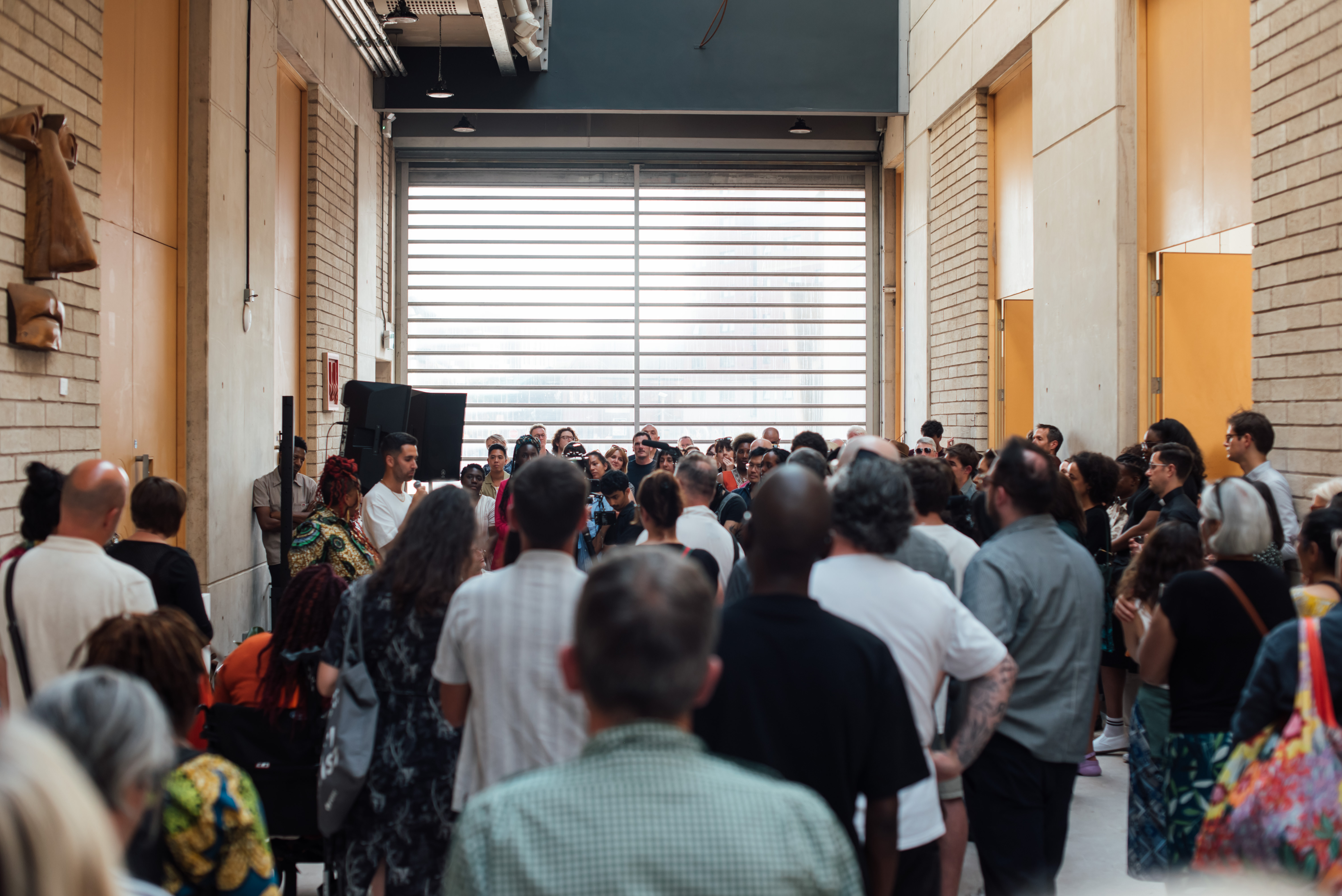 Busy crowd of people listening to a man speak on a microphone in a very tall atrium, doors to art studios line both sides.