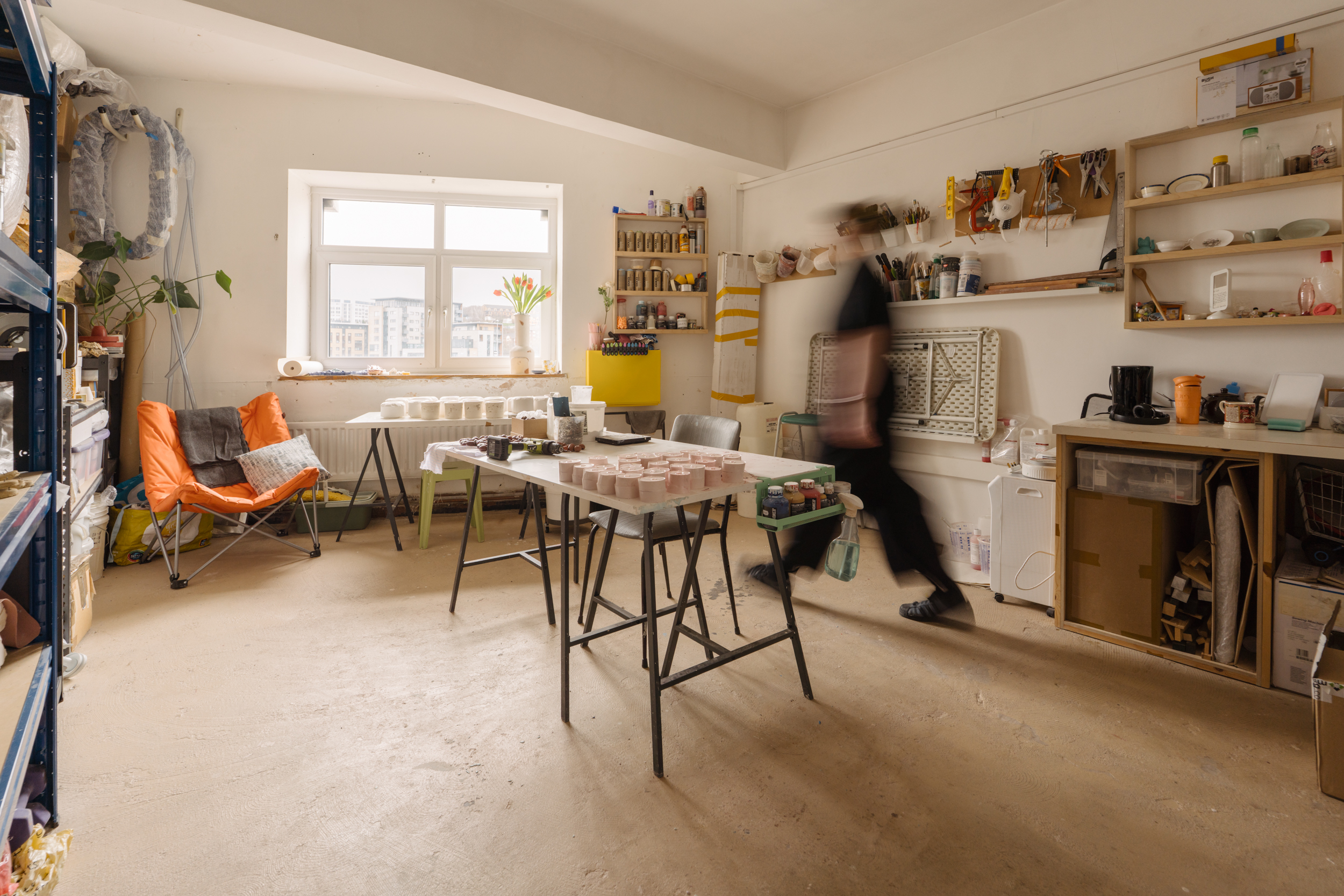 Spacious art studio with lots of wall storage, a workshop table set up in the middle, and a large orange chair in the corner. A blurred figure walks through.