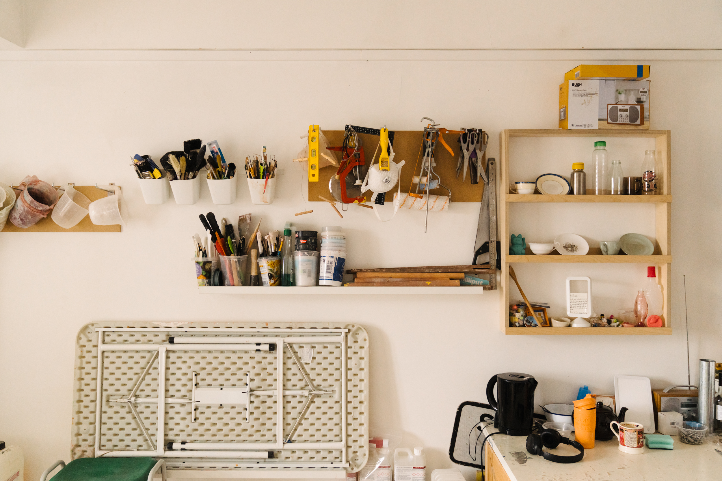 White studio wall with lots of storage for jugs, brushes, tools, facemasks, etc. Workshop table folded up and leaning against the wall. 