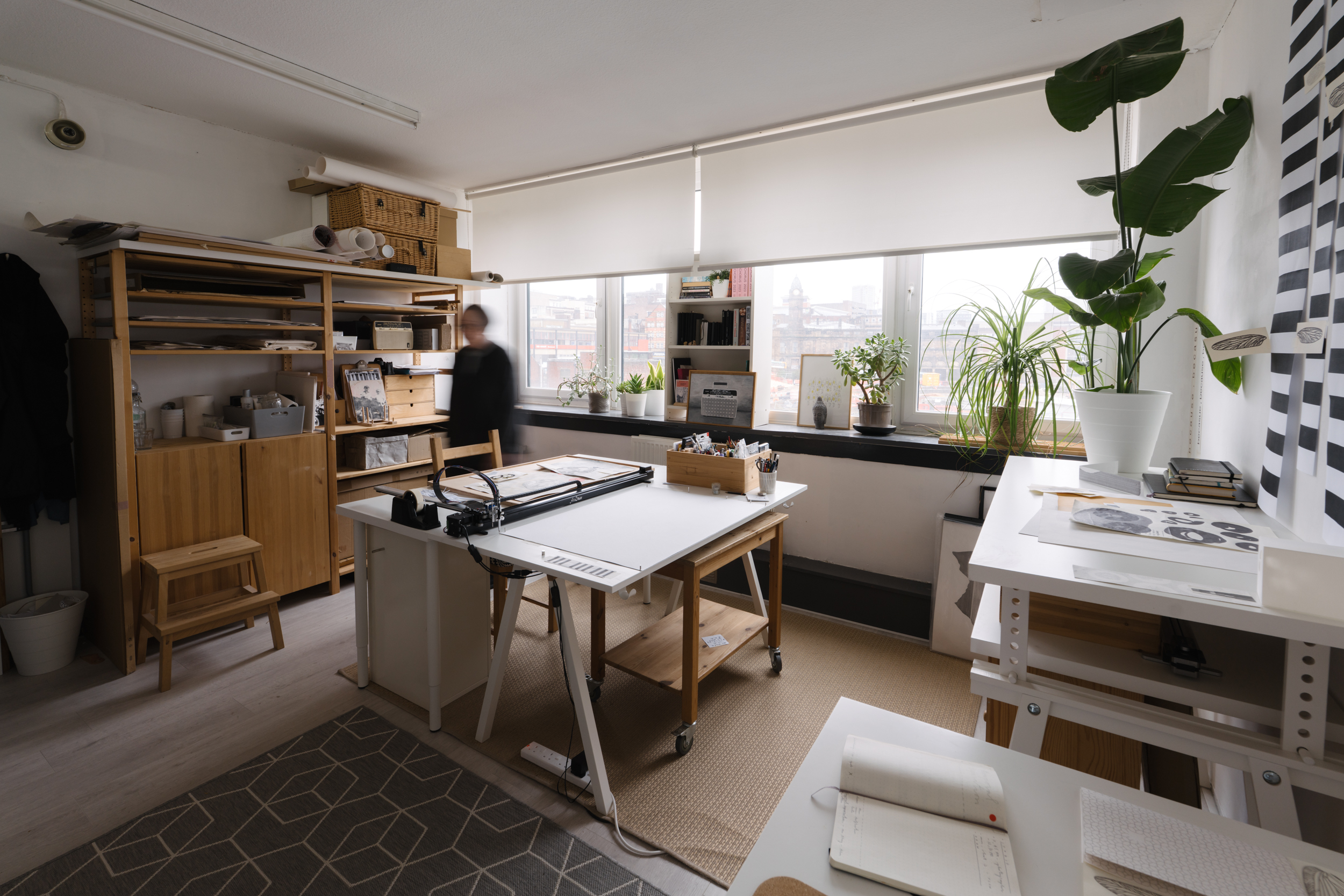 Spacious art studio with several desks set up for different kinds of work. Lots of plants, natural light through two wide windows. A blurred figure walks through.