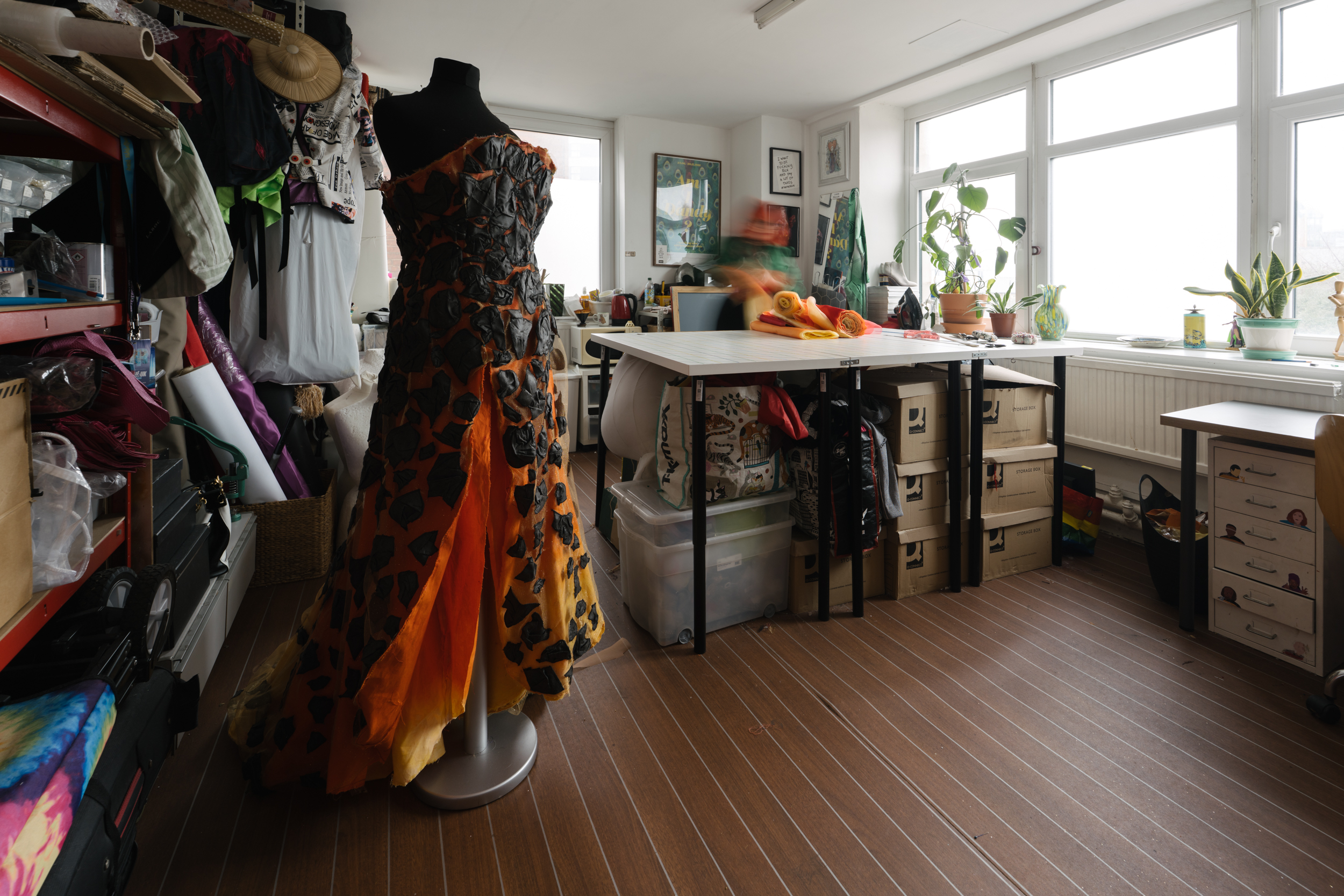 Spacious art studio full of materials and works in progress, including a dress on mannequin that looks like flowing lava. A blurred figure works at a large table.