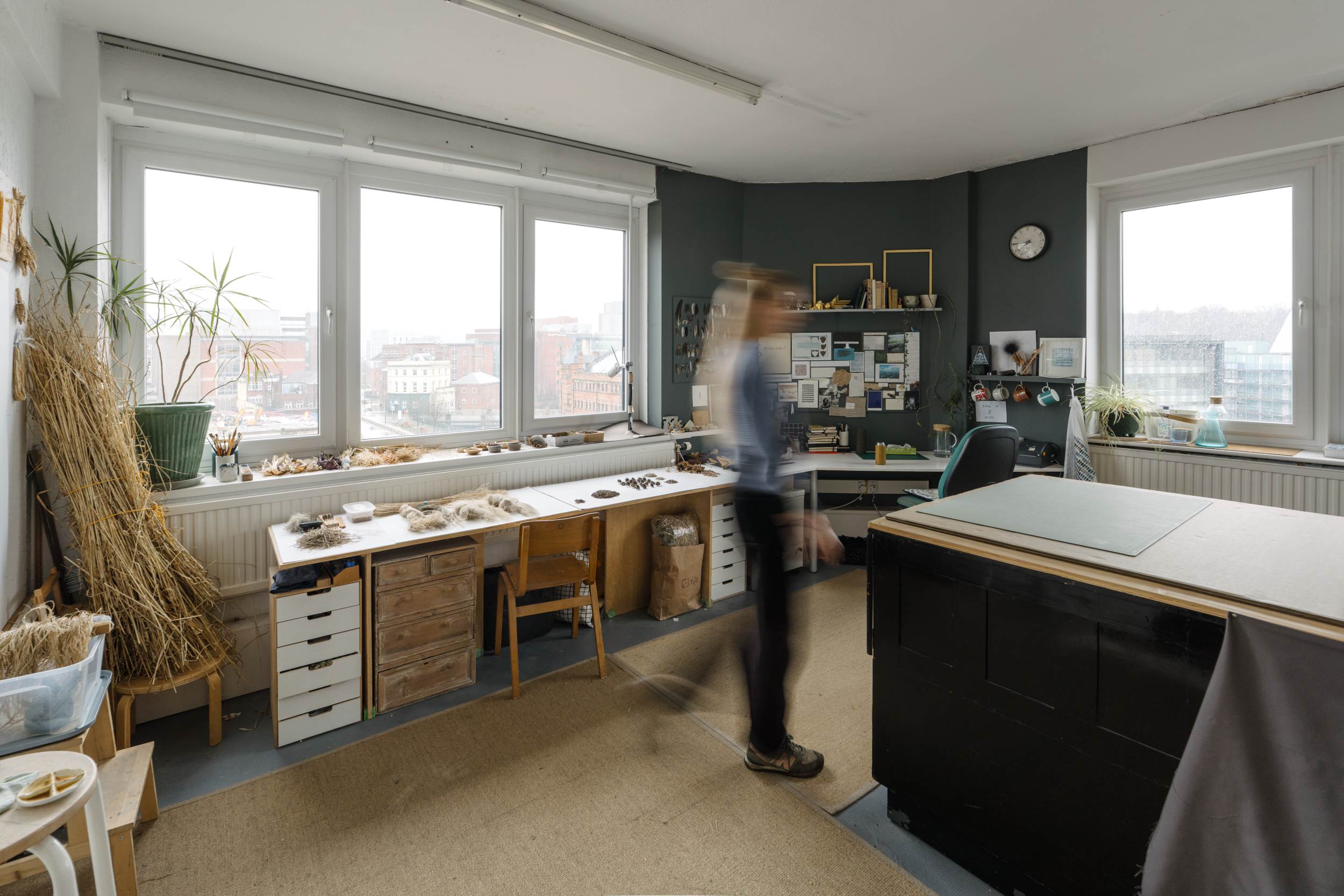 Spacious art studio with desks all along one side, lots of natural light, a large planchest, and a green wall with pictures and ephemera pinned up. A blurred figure walks through.