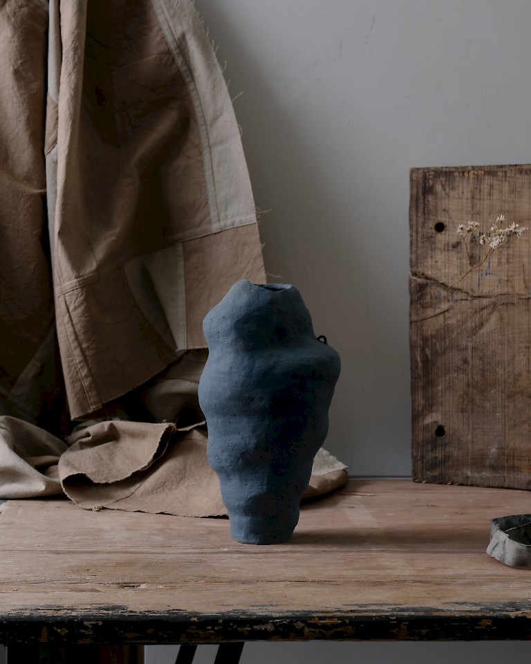 Wobbly black ceramic pot by George Baggaley, it looks like a plume of smoke with a tiny handle. Displayed on a workbench.