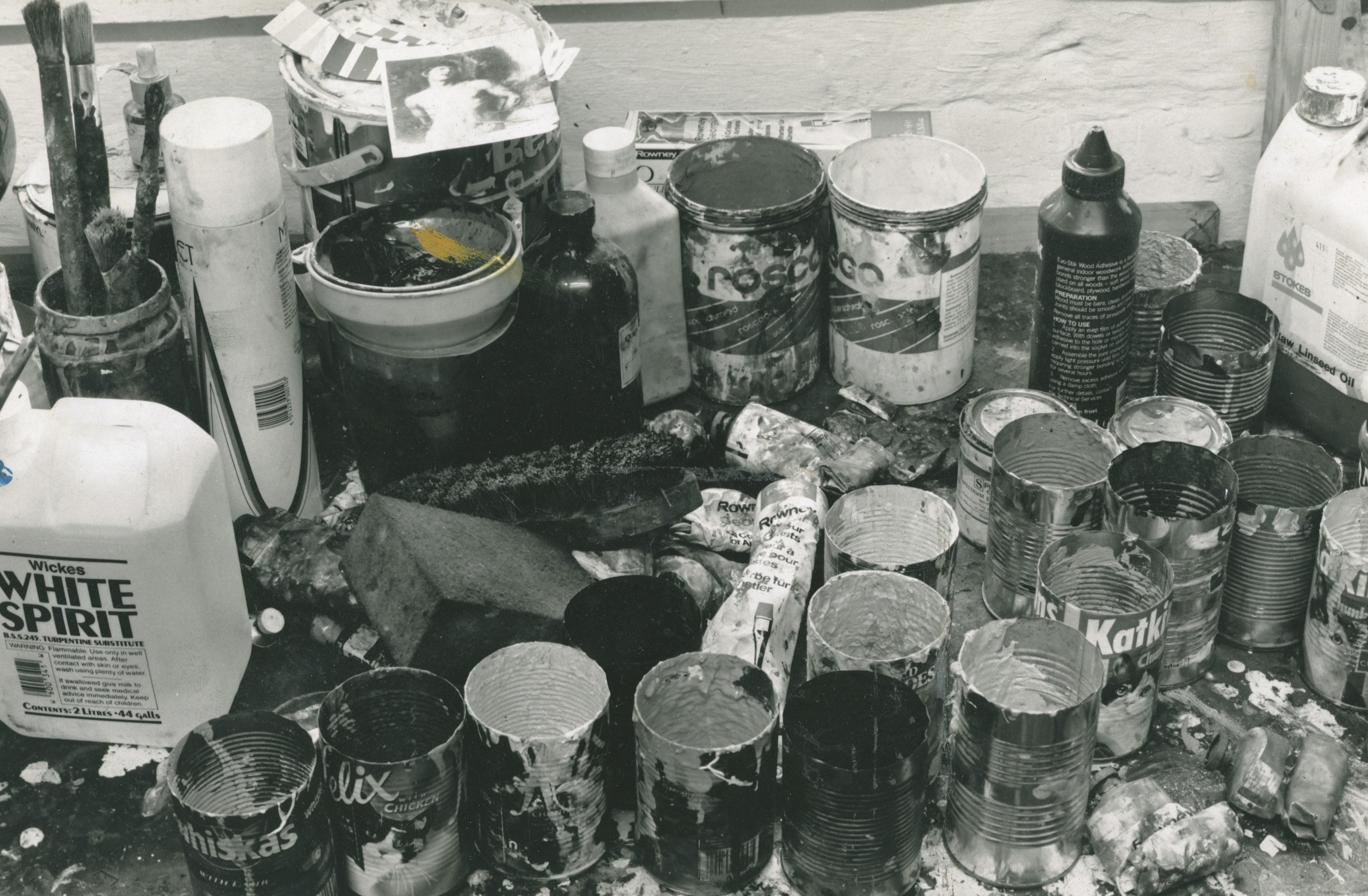 Black and white photograph of an artist's table, filled with pots of paint, white spirit and other materials.