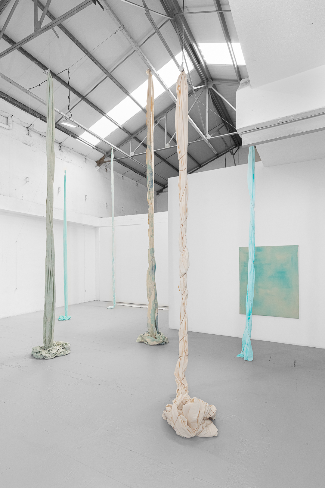 Installation by Jo Fleming Smith in a tall white gallery, lonf pieces of twisted blue fabric hang from the ceiling and bunch on the floor. Blue canvas in the background.