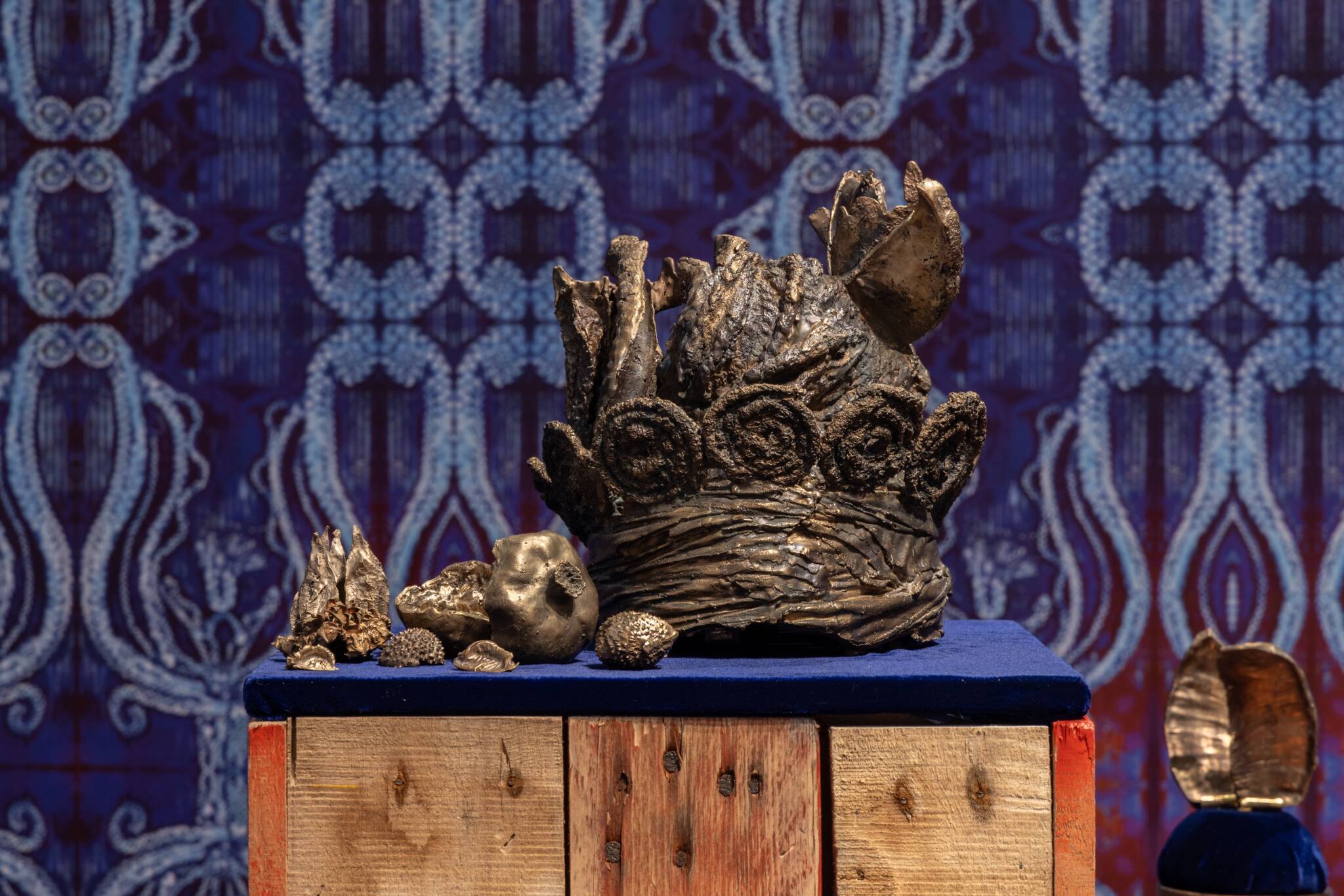 Bronze sculptures of a braided headdress and small fruits by Kedisha Coakley, displayed against a royal blue wallpaper background.