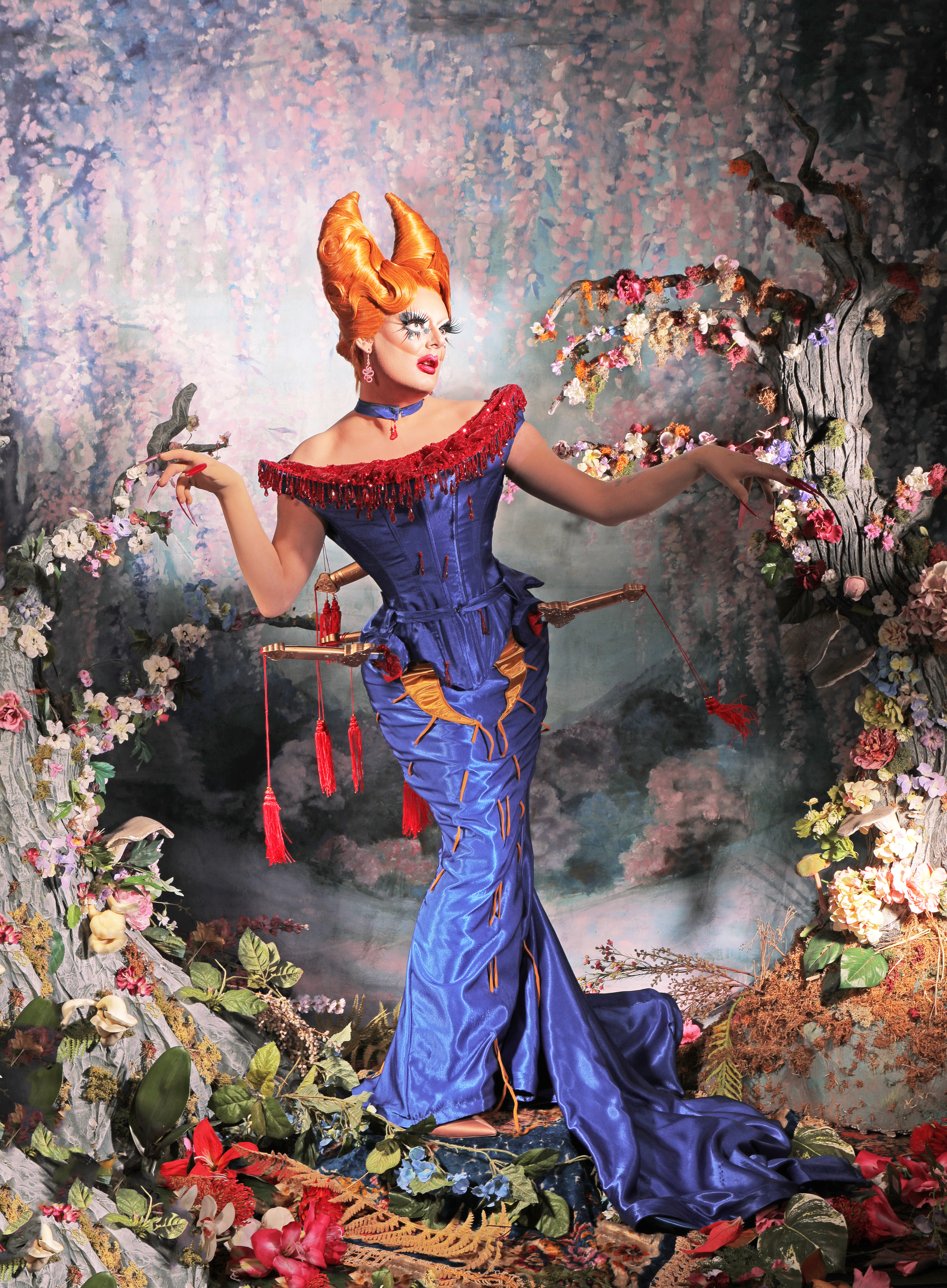 Drag queen wearing a blue satin gown by Lizzie Biscuits, there is a beaded dripping blood effect at the top and several gold swords with red tassles appear to be stabbing into the material.