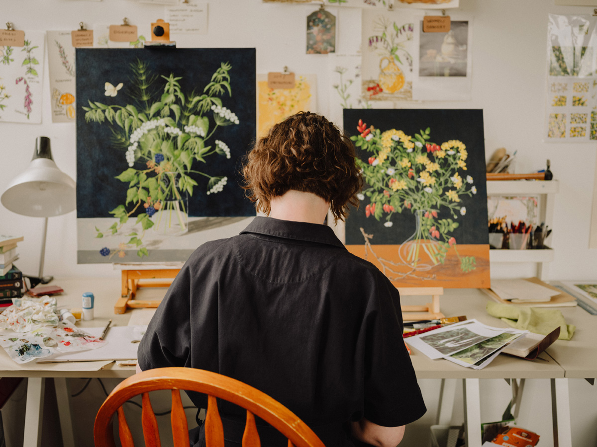 Woman with a brown bob and a black shirt with her back to the camera, sat at a desk with two unfinished still life paintings. On the wall and desks are lots of sketches and reference images.