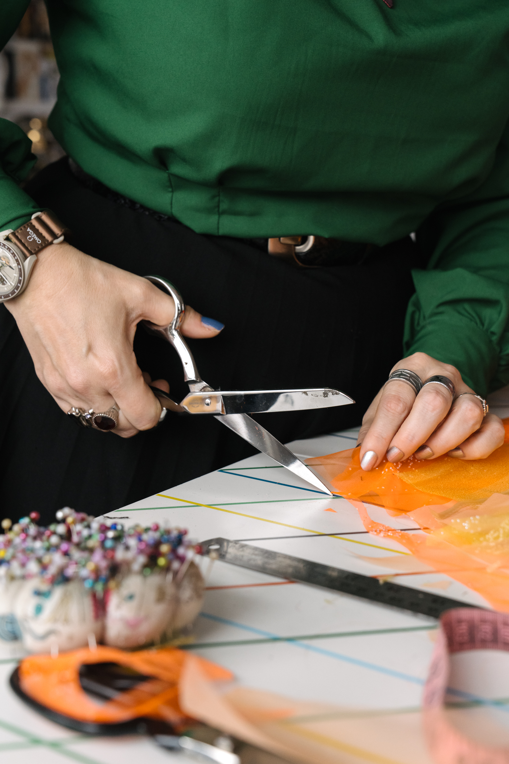 Woman in emerald green blouse, blue and gold nail polish, and lots of jewellery using a pair of silver scissors to cut through some orange tulle fabric.