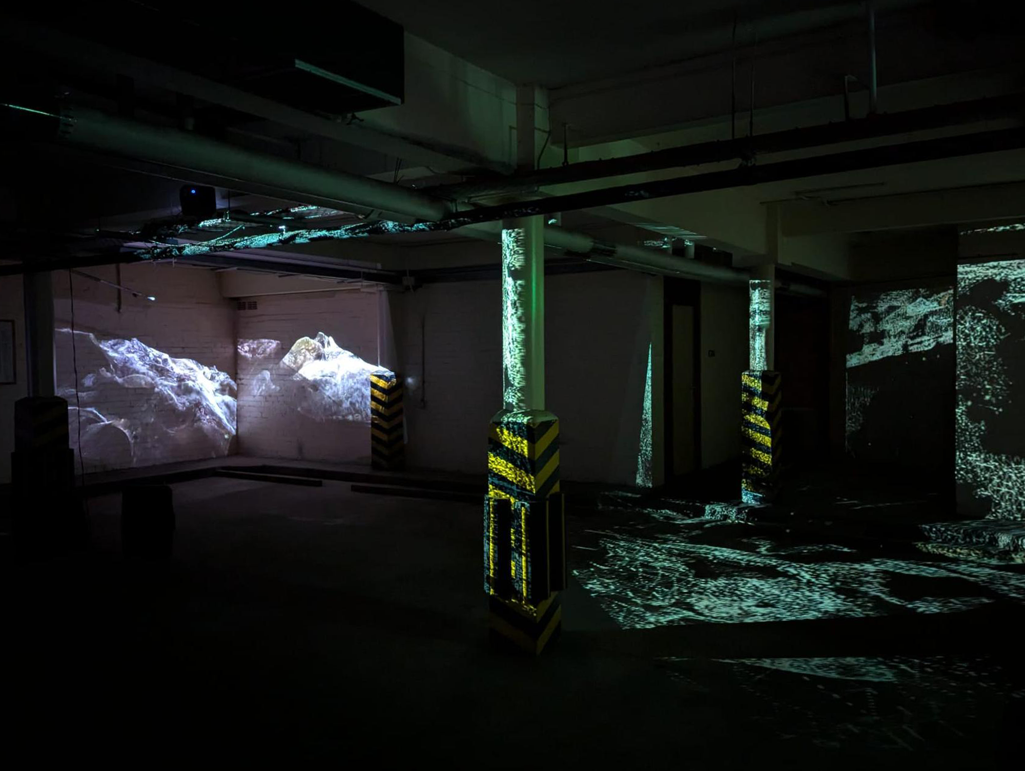 Dark underground space with overhead pipework and supporting pillars with yellow and black hazard stripes; various projections of watery digital imagery on the walls and floor.