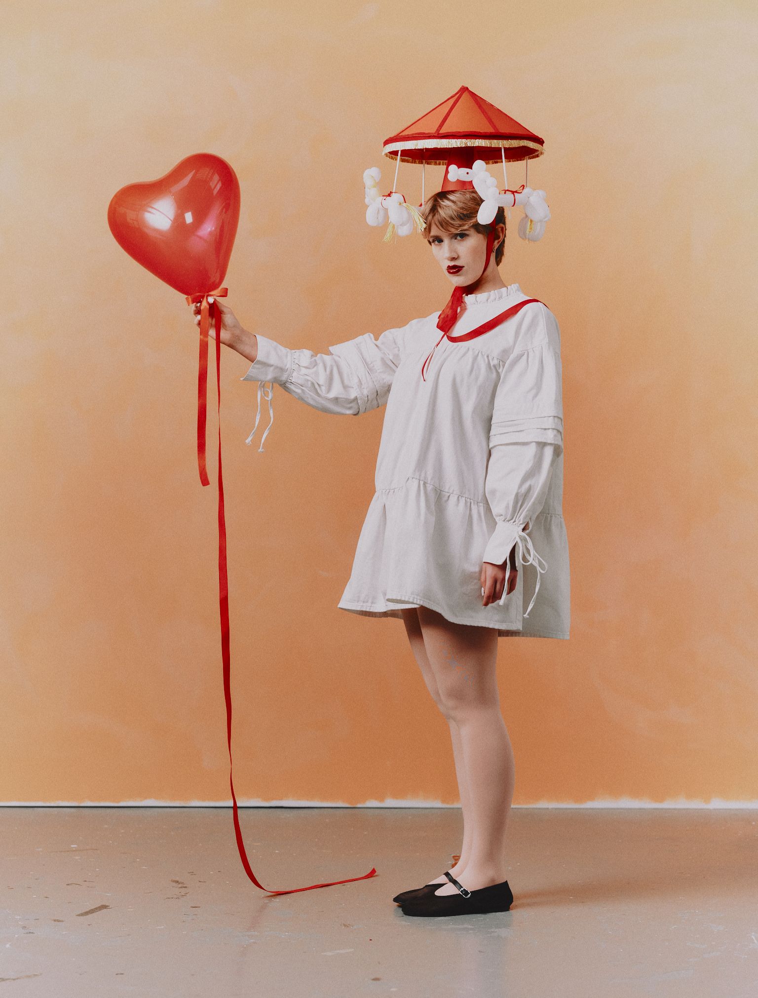 Photograph by Samuel Binstead. A woman in a white smock dress holding a red heart-shaped balloon, wearing a whimsical hat that looks like a merry-go-round with balloon animals hanging from it.