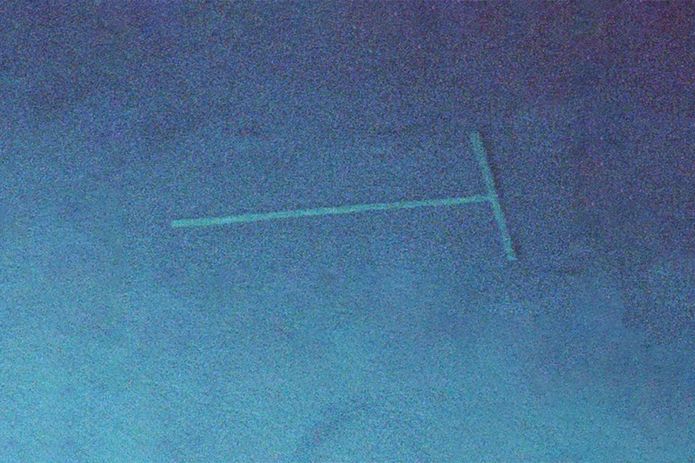 A pin or other T-shaped object on its side. It seems to be underwater, surrounded by deep blue. The image quality is grainy.