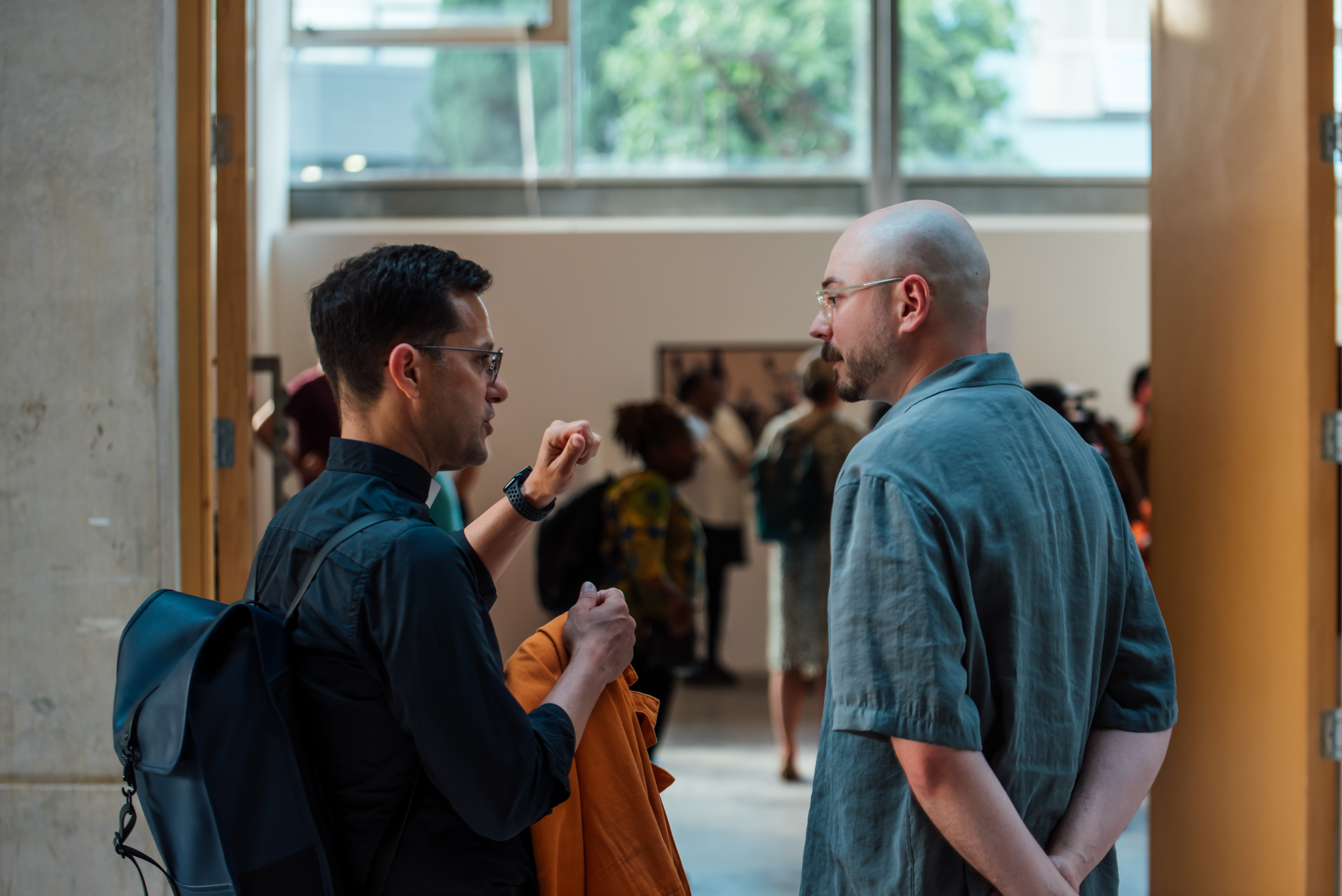 Two men chatting at an art event, a crowd of people looking at art in the background.
