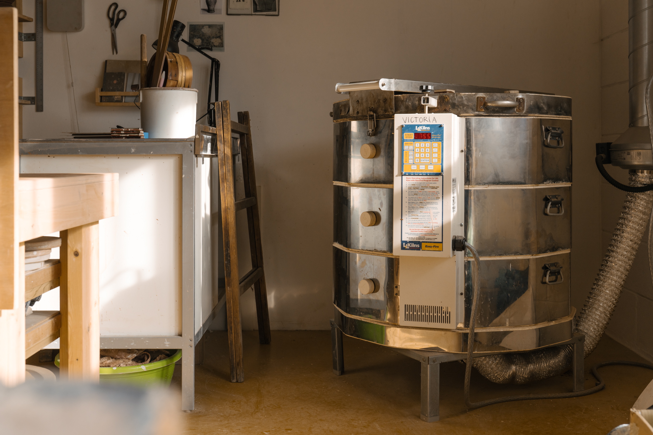 Pottery studio with large round silver kiln, the name 'VICTORIA' written across the control panel in black marker.