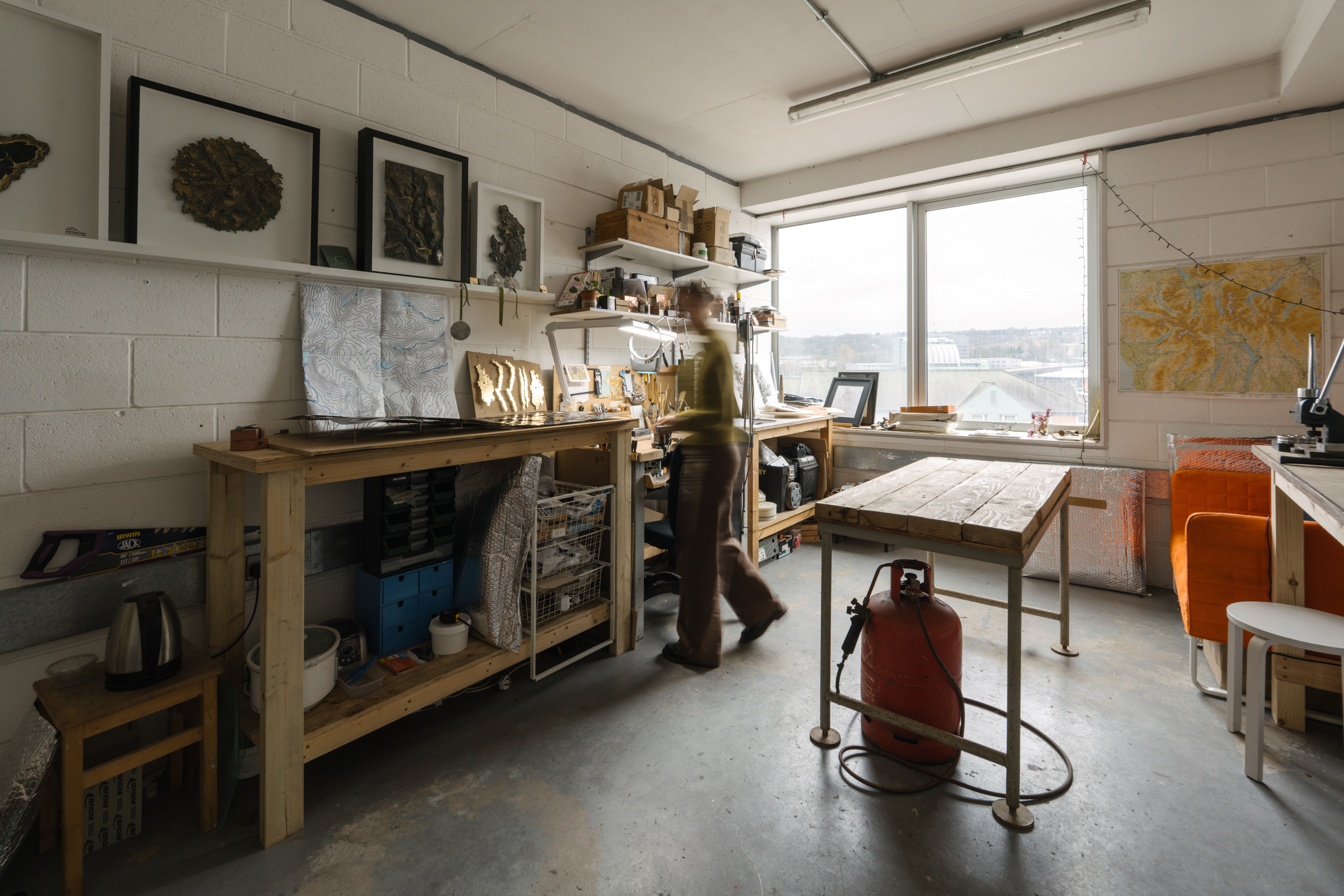 Spacious art studio with views over the city behind. A long jewellery bench on one side and a blurred figure working.