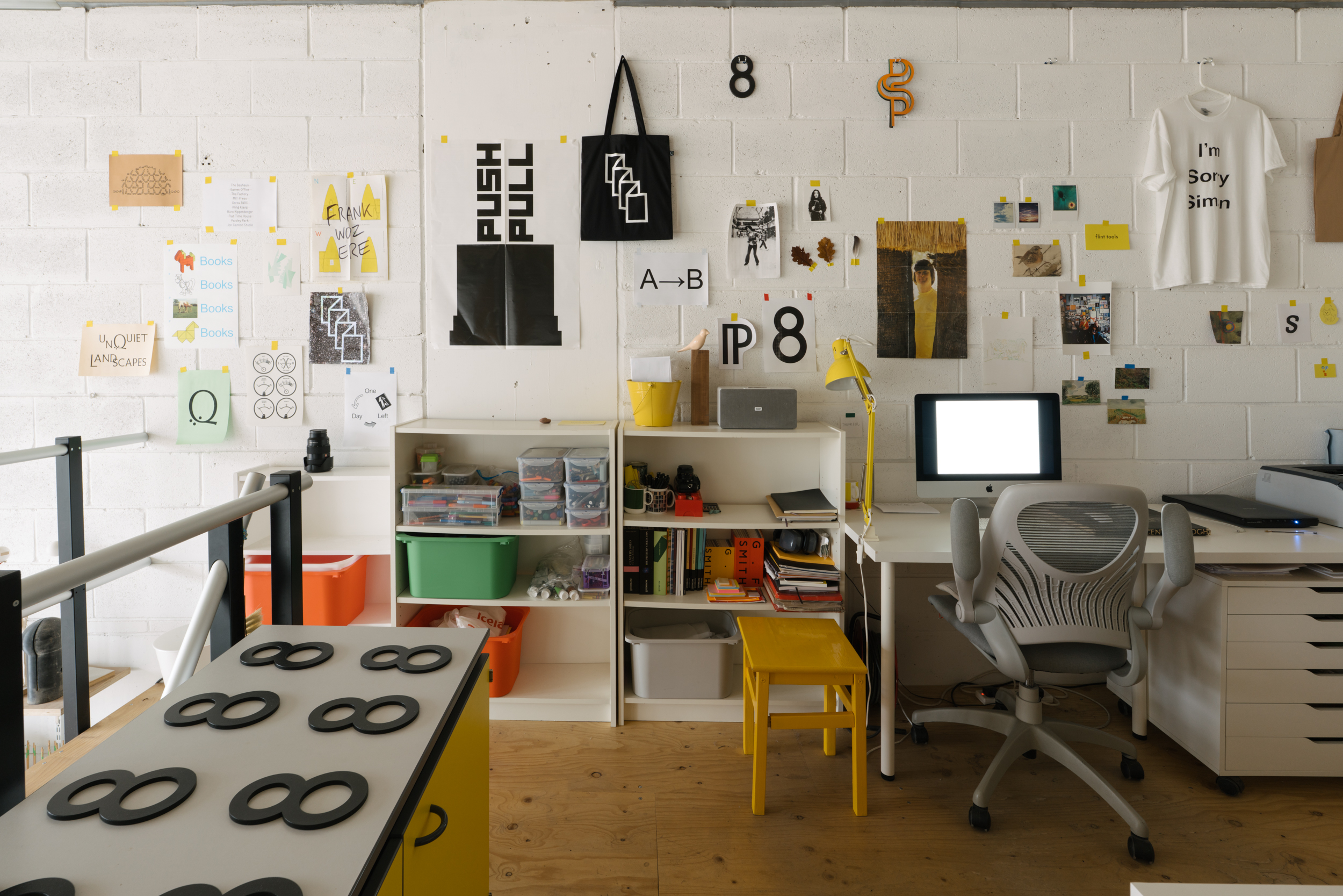 Bright studio with white breezeblock walls, showcasing a range of works on paper and a tshirt reading 'I'm Sorry Simon'. Desk with computer.