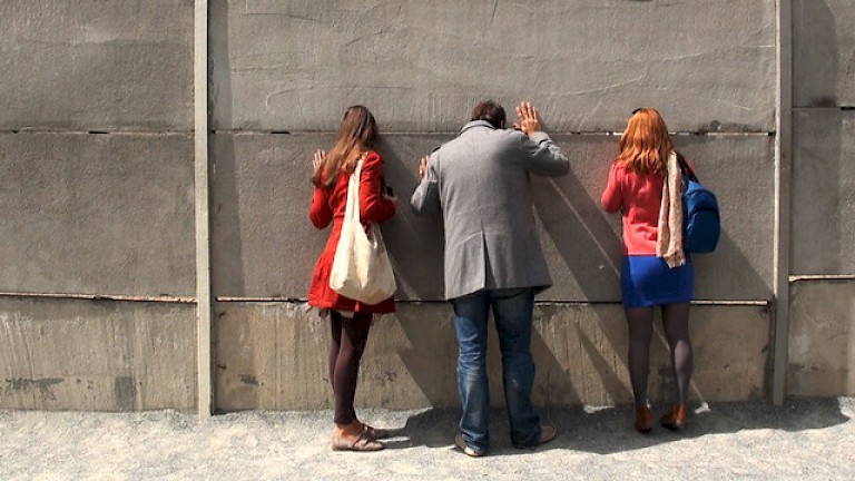 Photograph by Rose Butler; three people with their backs to the camera peer through a small gap in a concrete wall.