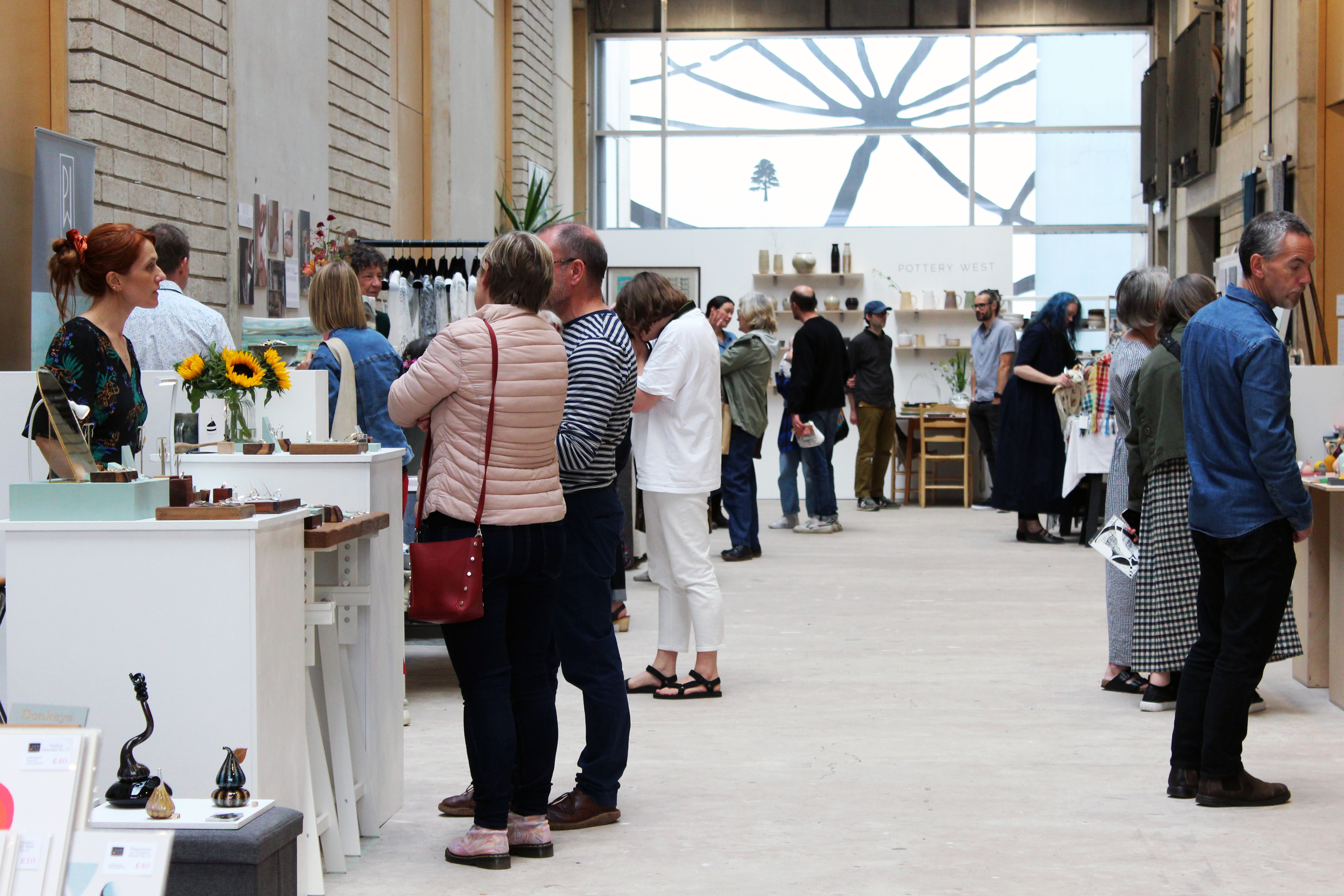 Tall concrete atrium with lots of light, hosting an art fair with many artists stood at stalls of their work on both sides, talking to customers.
