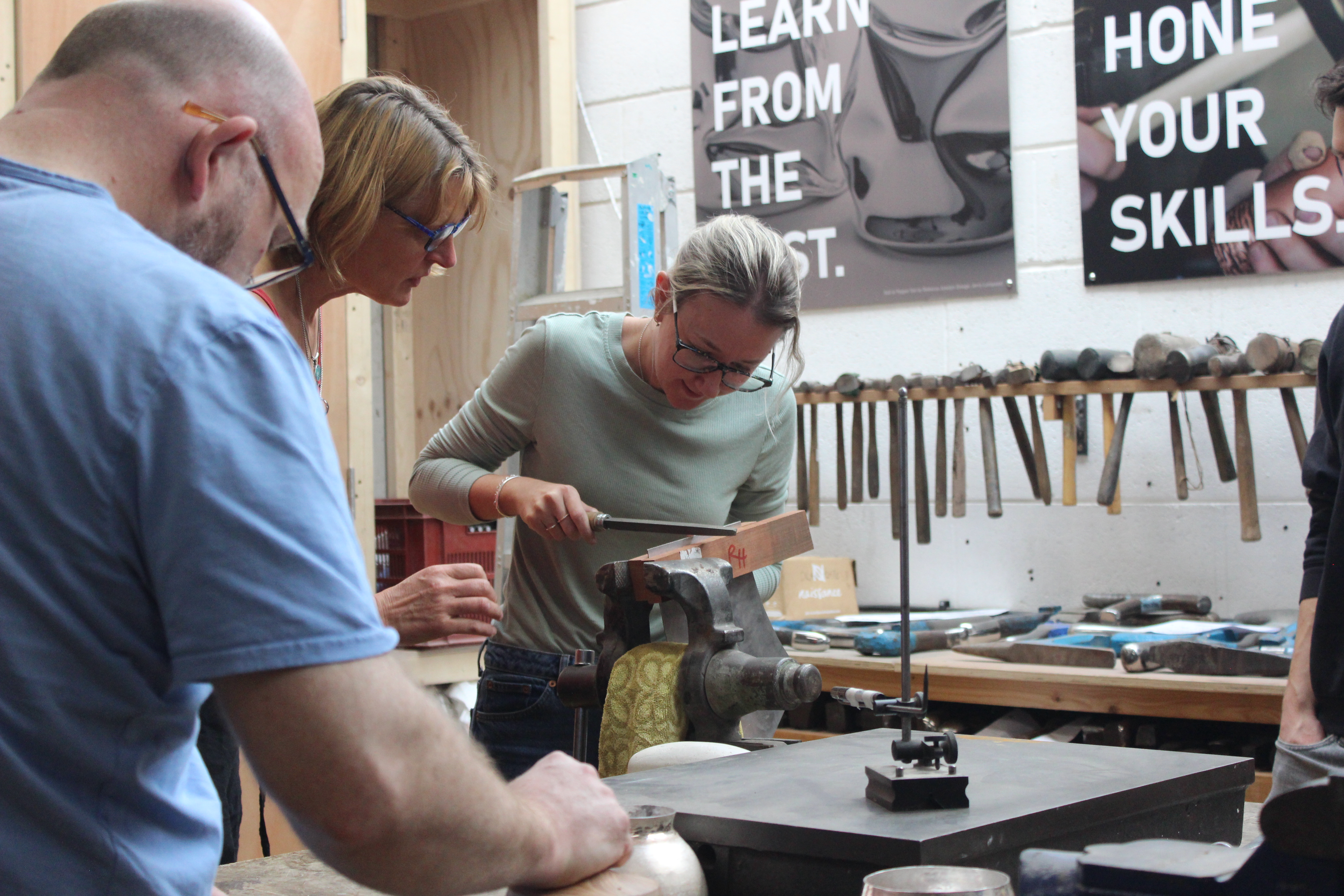 Young blonde woman in green shirt filing metal in a vice, while another blonde woman supervises. 