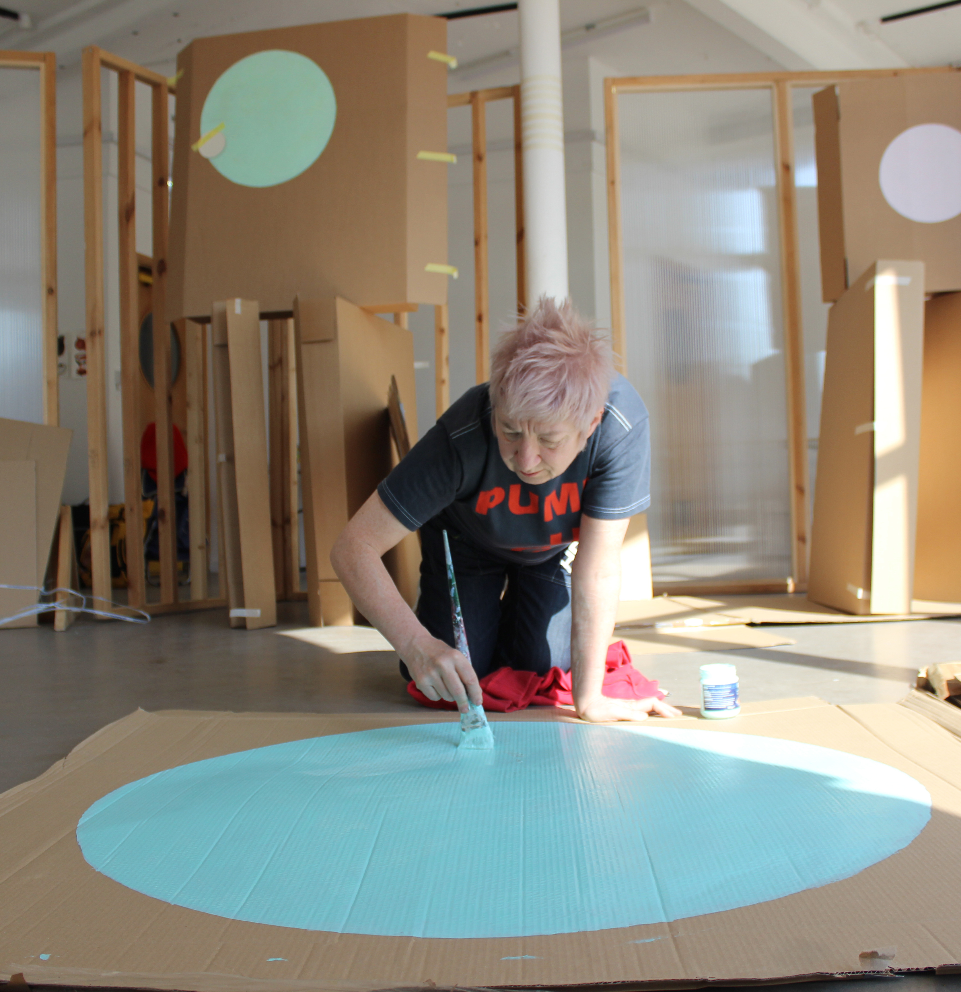 An artist with short pink hair painting a blue circle on a large piece of cardboard, behind them is more cardboard with different coloured circles.