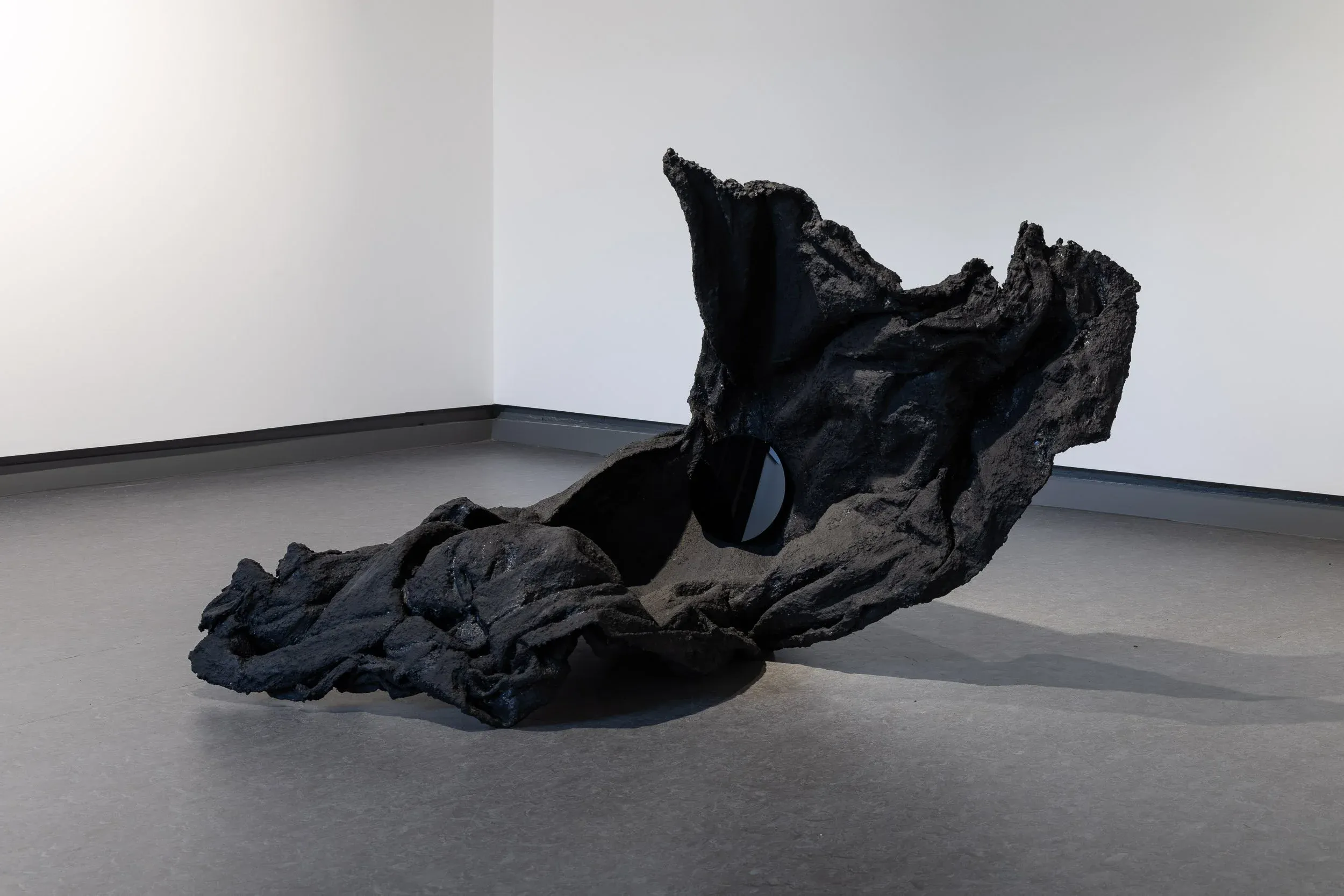 Black fabric hardened into a solid form that lifts off the ground, looks like stone or part of a cliffside. A small round black mirror sits within a crevice.