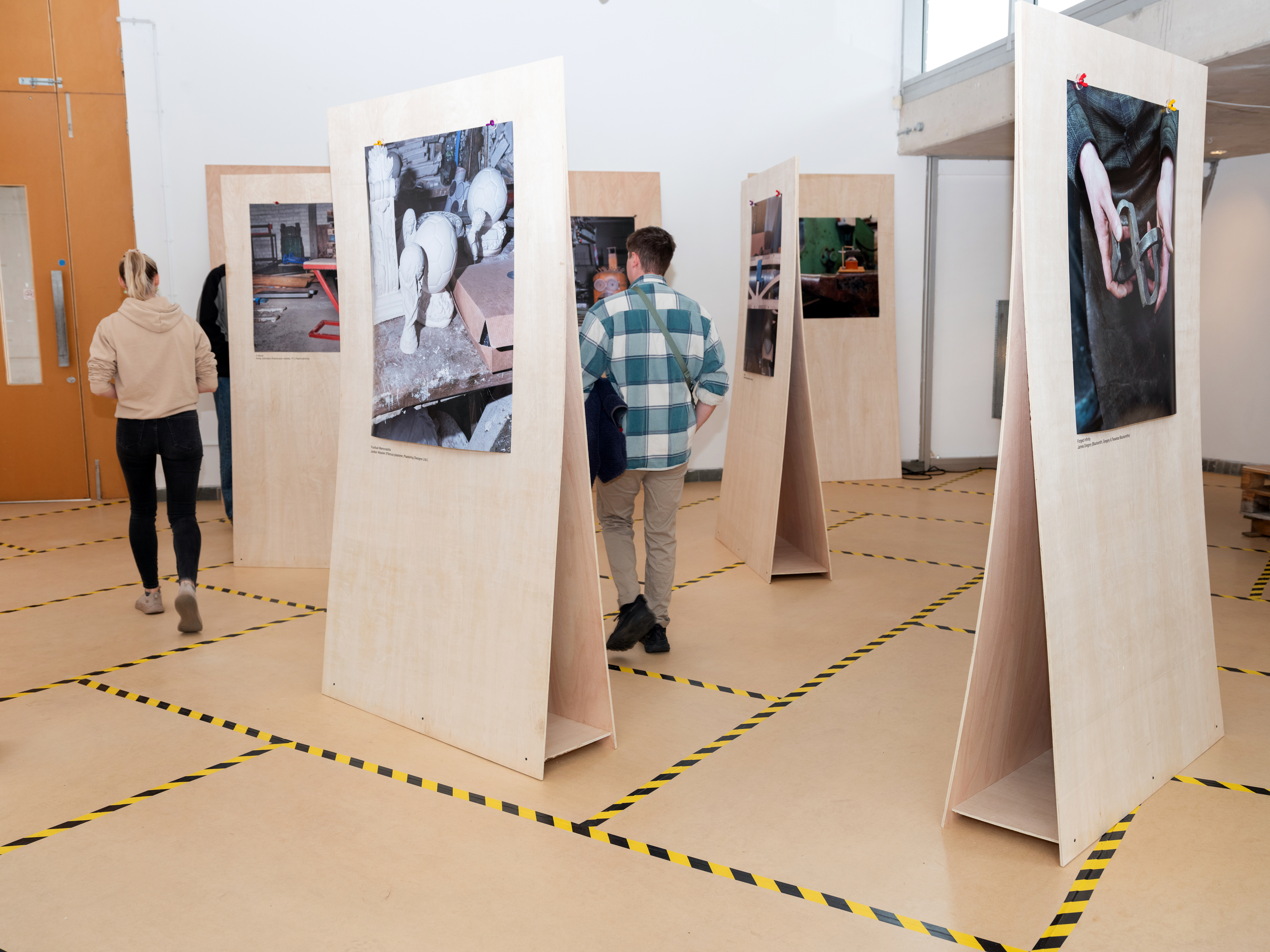 Installation view of a photography exhibition, images displayed on freestanding plywood sheets and floor covered in plywood held together with black and yellow striped tape.
