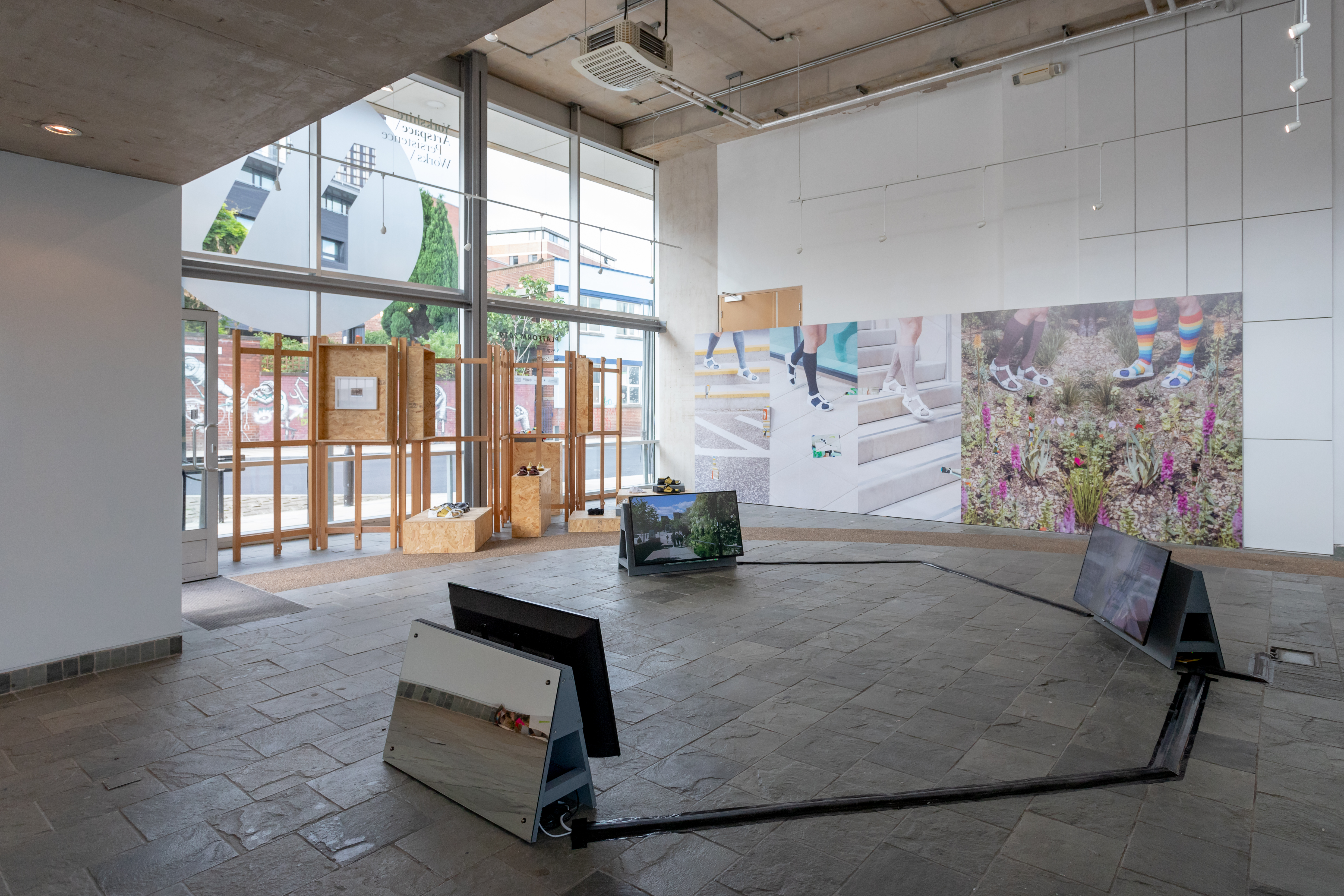 Installation view of three screens displayed on the floor, in a semi-circle. Colourful photographs of feet wearing sandals in the background.
